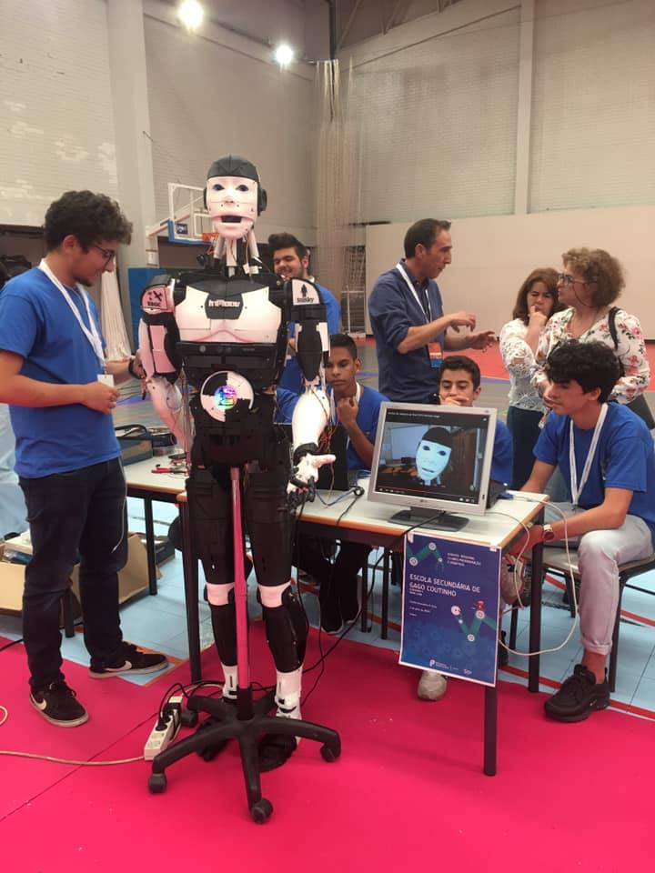 The robot on its stand at a competition venue, surrounded by team members in blue shirts, with its camera feed shown on a monitor.