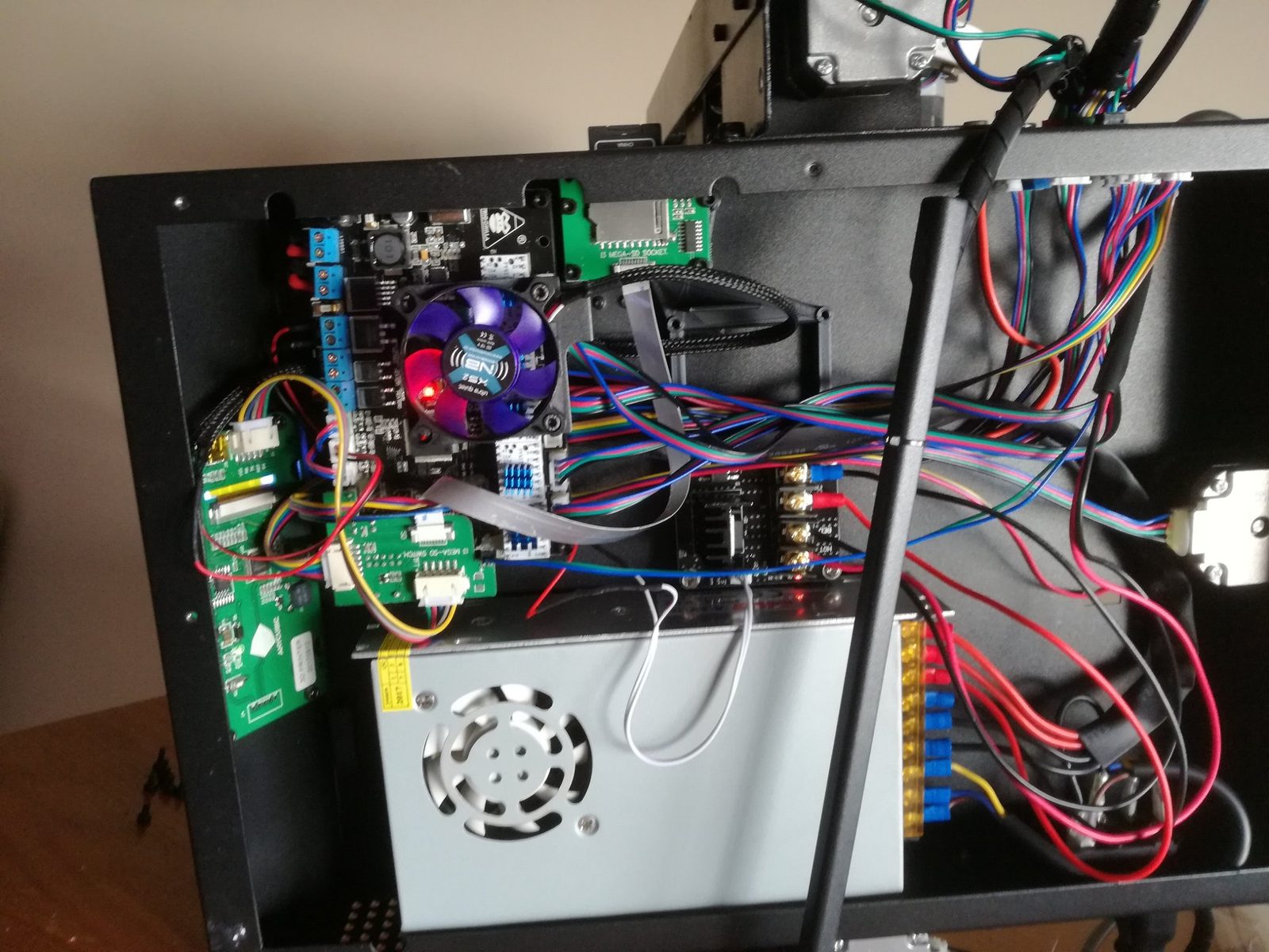 The open underside of a 3D printer showing a rewired mainboard, cooling fan, power supply and colour-coded cabling.